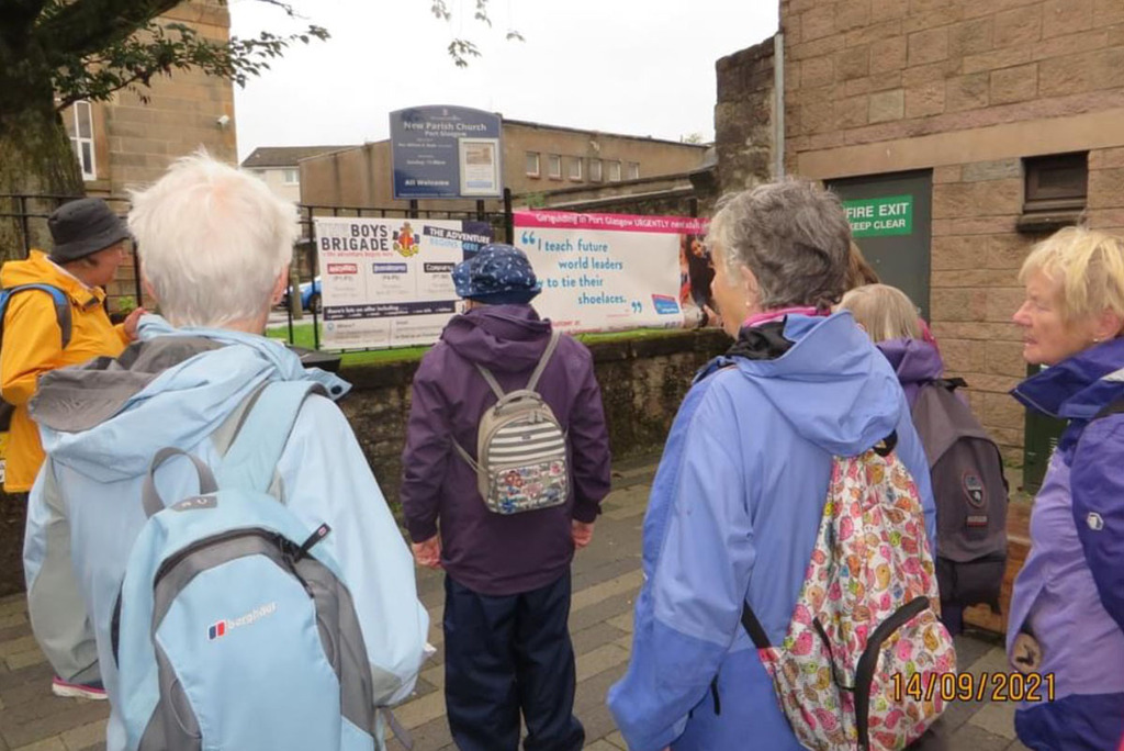 Six women all look towards a Girlguiding recruitment sign. The women are all wearing jackets and backpacks of different colours. They are looking at the recruitment banner that is on railings.