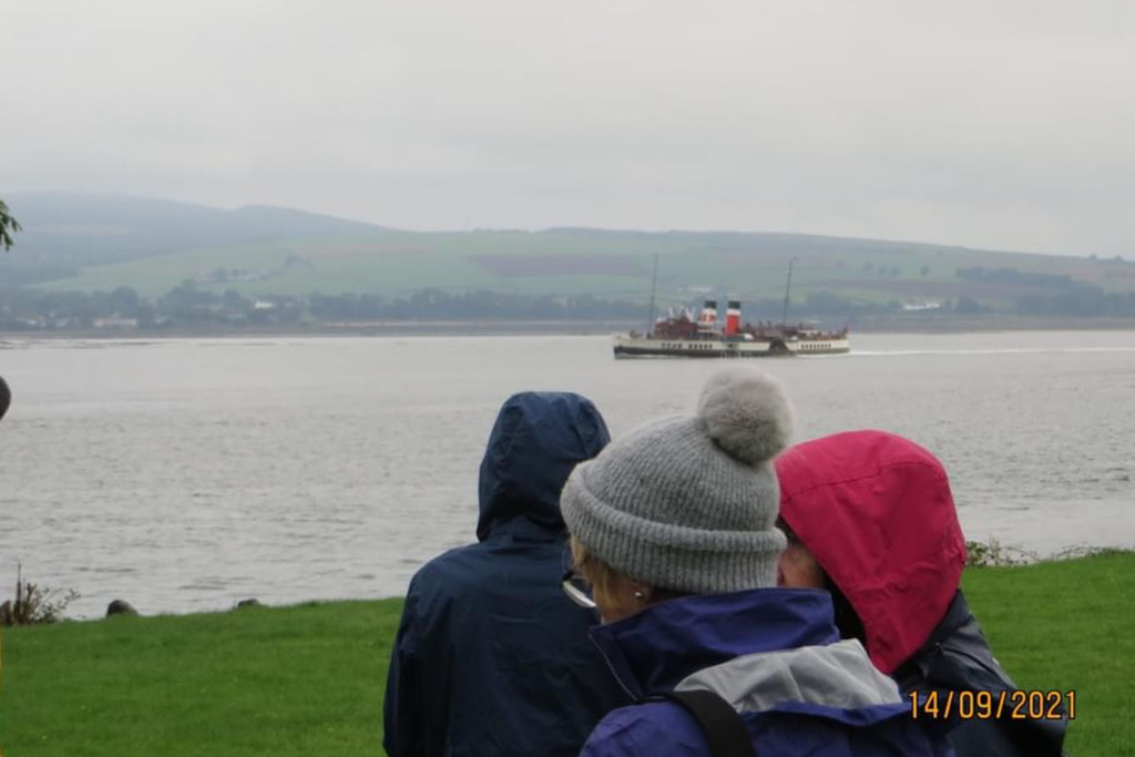 Three women stand in the foreground, and in the middleground is the Waverly: a paddle steamer. The sky is grey.